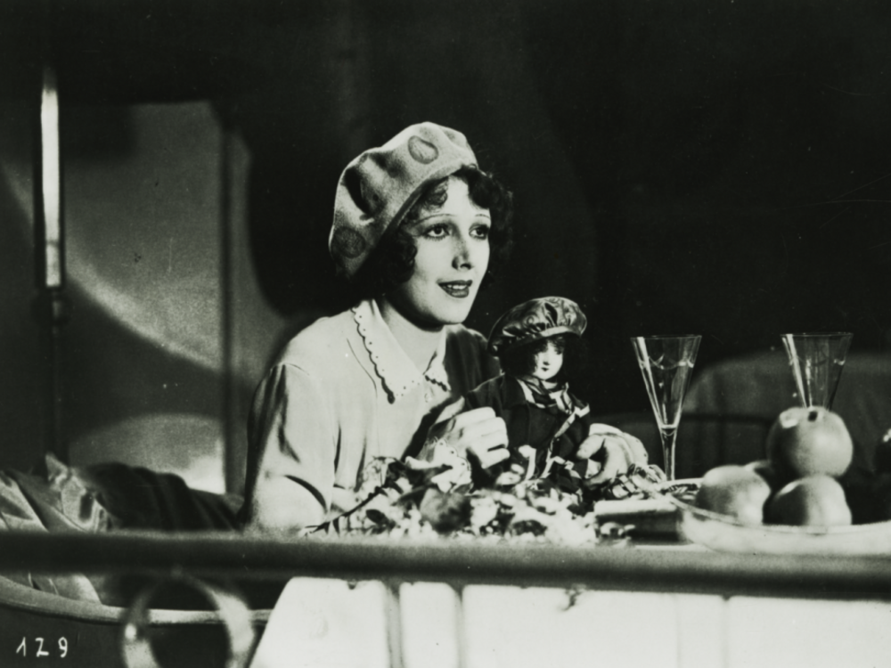 Black and white image from the movie The Convict from Stambul with a woman at the table holding a doll in her hand.