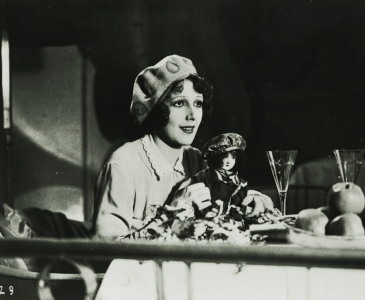 Black and white image from the movie The Convict from Stambul with a woman at the table holding a doll in her hand.
