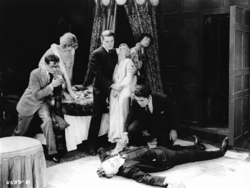 A group of people in 1920s attire is looking at the floor, where a man, seemingly murdered, lies. 