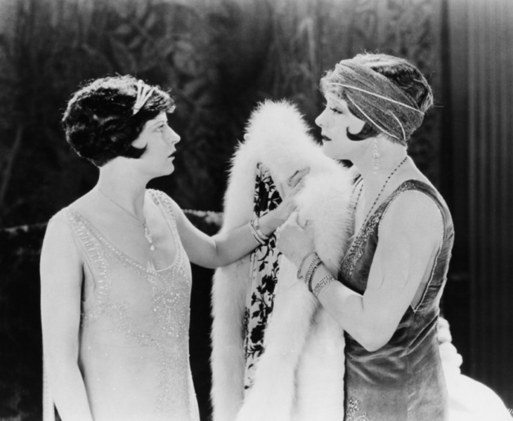 Two women in 1920s clothing and hairstyles are facing each other. One is holding a white fur.