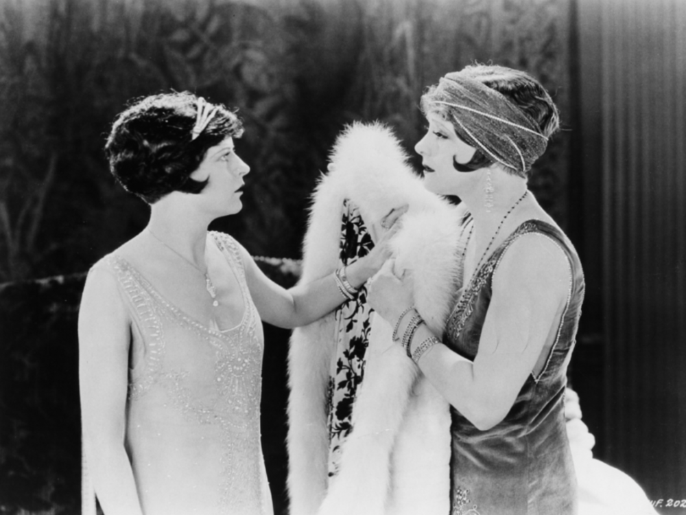 Two women in 1920s clothing and hairstyles are facing each other. One is holding a white fur.